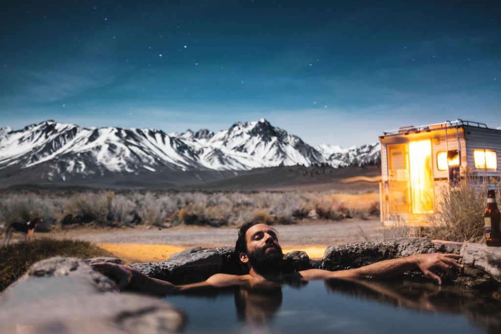 man relaxing in natural spring with truck camper and snow topped mountains in the background